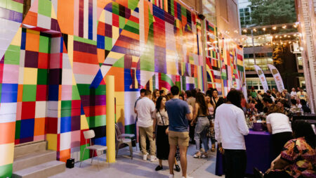 A vibrant mural in bold colors adorns the wall as attendees gather at a lively outdoor event, with tables and string lights illuminating the scene.