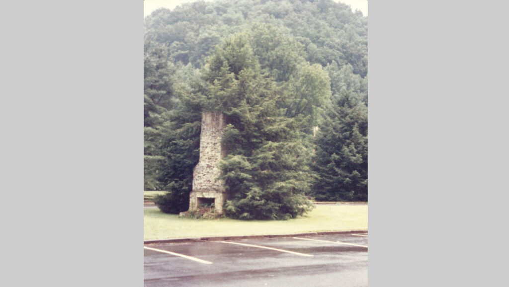 A stone chimney partially engulfed by greenery, surrounded by a grassy area and trees, with a mountain backdrop.