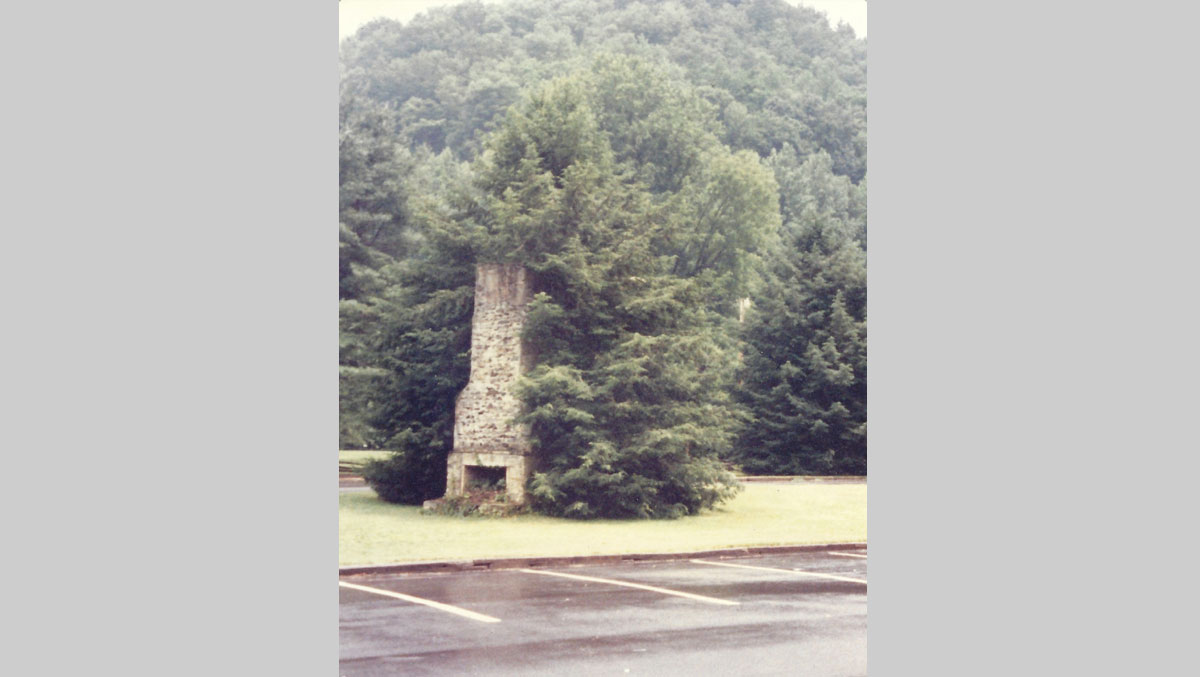 A stone chimney partially engulfed by greenery, surrounded by a grassy area and trees, with a mountain backdrop.