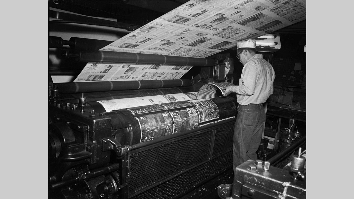 A man operates a vintage printing press, overseeing newspaper production as printed sheets roll out above him in a dimly lit factory.