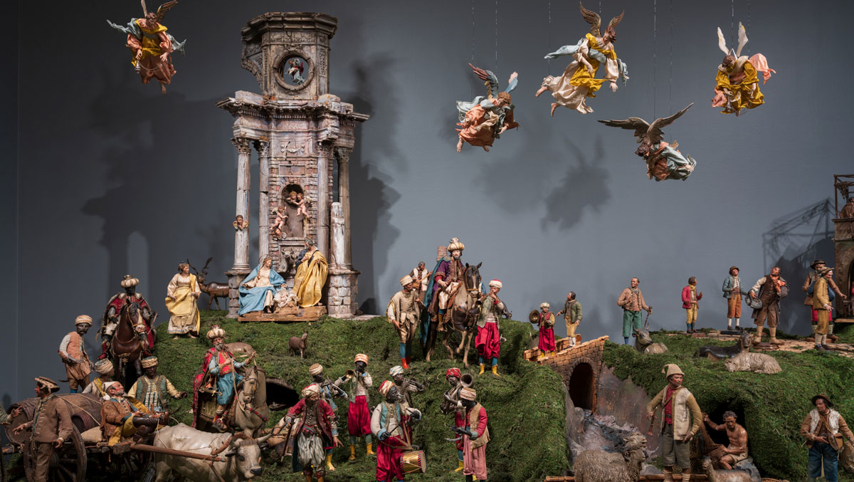 A detailed nativity scene features numerous figures in traditional attire, lush greenery, and a backdrop of ornate architecture and angels.