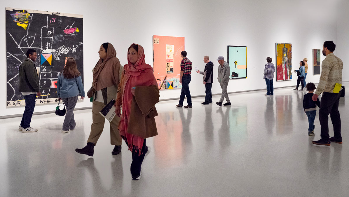 A busy art gallery featuring multiple paintings on the walls, with visitors walking and engaging with the artwork.