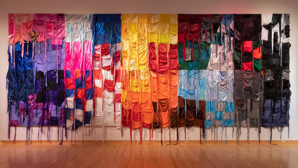 A vibrant textile mural composed of draped colorful fabrics, creating a flowing, abstract pattern on a gallery wall.