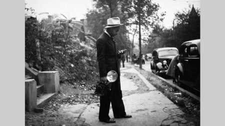 A black & white photo of a man in a fedora standing on a sidewalk, holding a vintage camera and light, surrounded by trees and classic cars.
