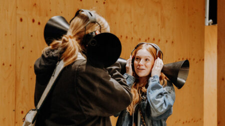 Two individuals engage in a playful sound experiment, each holding a large black cone to their ears against a wooden backdrop.