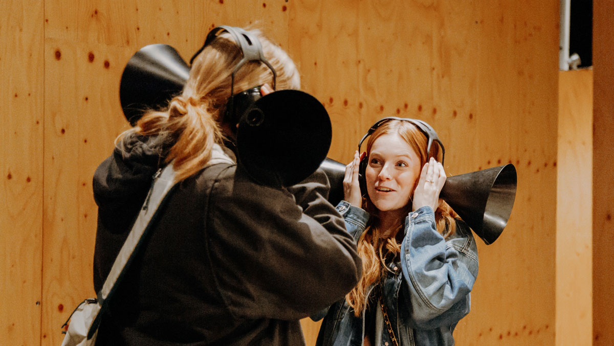 Two individuals engage in a playful sound experiment, each holding a large black cone to their ears against a wooden backdrop.