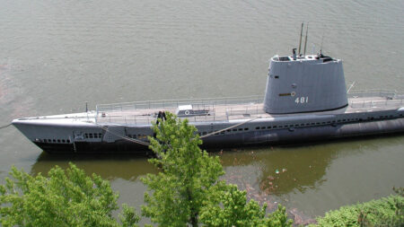 A vintage submarine, numbered 481, is docked on a river, partially obscured by trees, showcasing its sleek metal design.