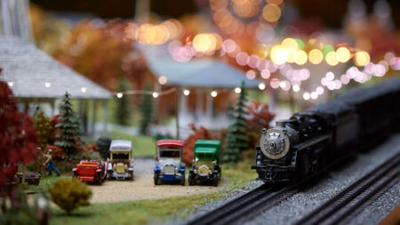 A miniature train engine travels past vintage toy cars and a festive, brightly lit backdrop of fall foliage and charming structures.