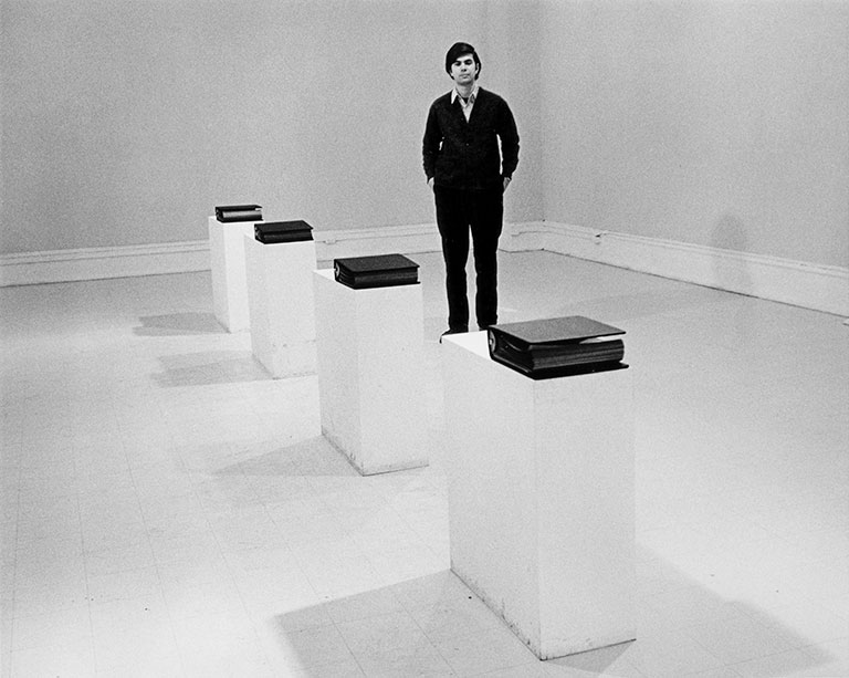 Photo of the artist from the 1960s, standing in a gallery surrounded by 4 tall pedestals each topped with a thick black binder.