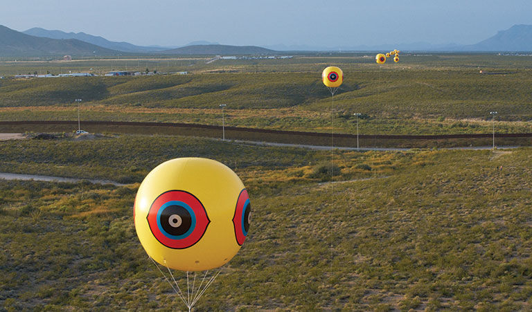 weather ballons hovering over a brown landscape. The balloons have eys painted on them.