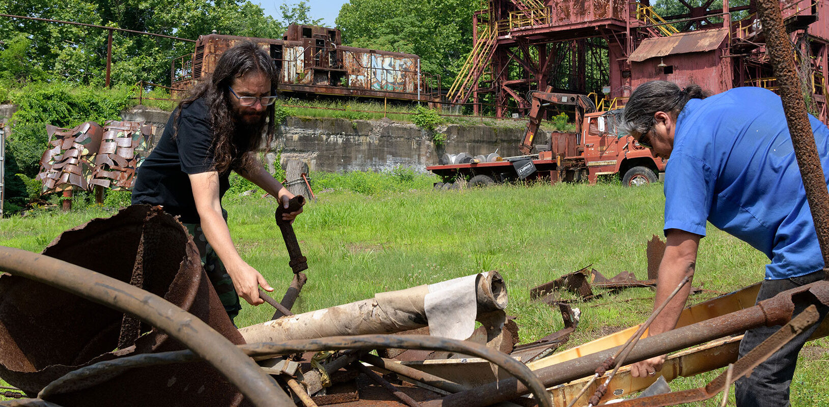 Two men sorting through rusted scrap metal in front of a rusted abandoned steel mill.