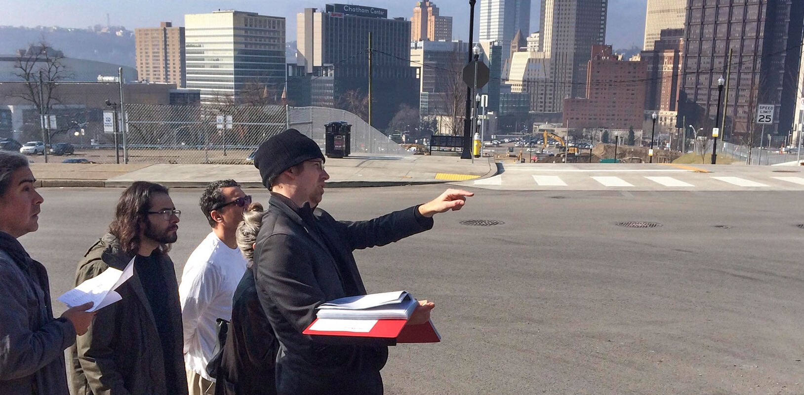 Group of people standing together with the city of Pittsburgh behind them. One man is pointing and holding a book.