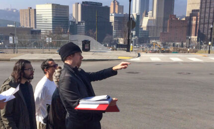 Group of people standing together with the city of Pittsburgh behind them. One man is pointing and holding a book.