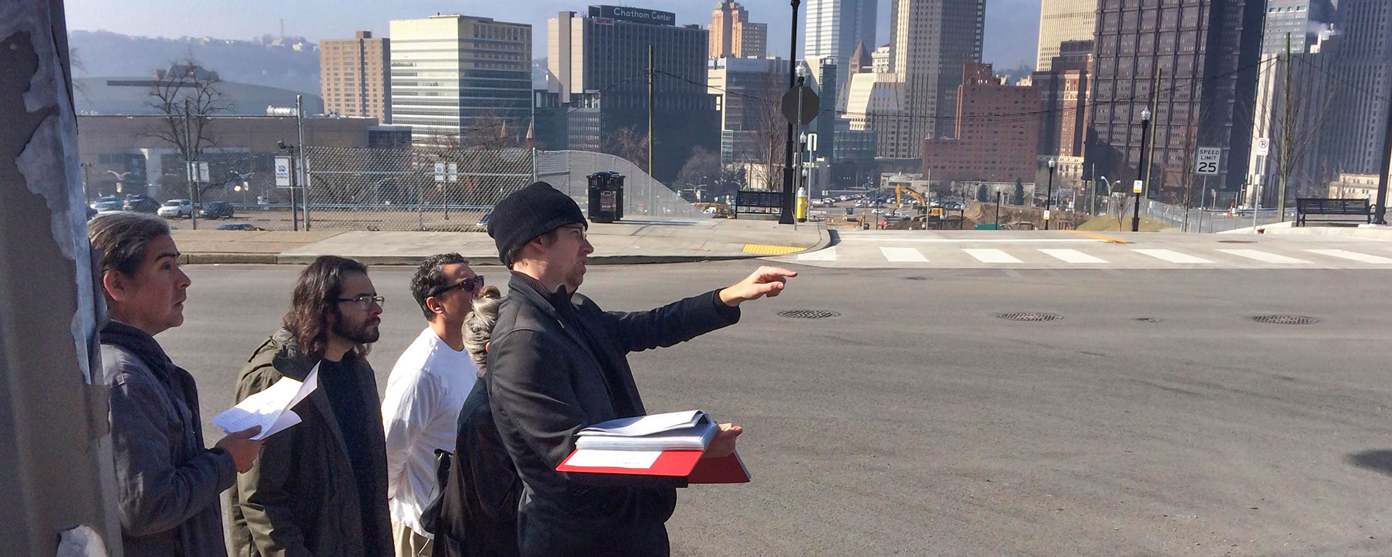 Group of people standing together with the city of Pittsburgh behind them. One man is pointing and holding a book.