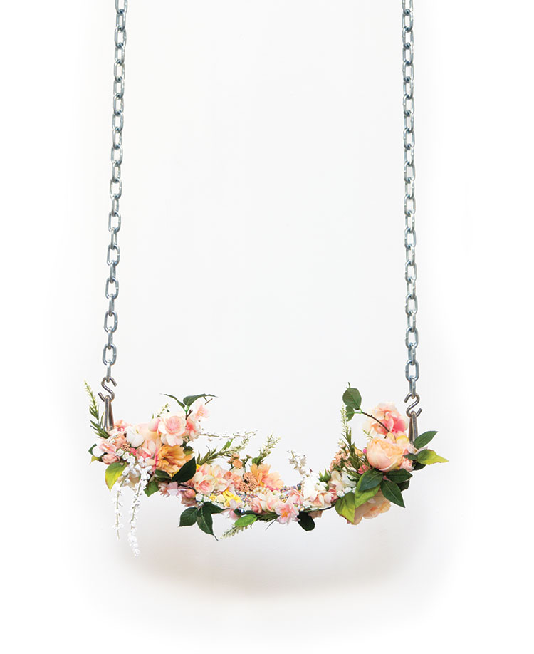 A sculptural artwork depicting a swing with aluminum chain suspension and a seat of peach, pink, white and yellow flowers. The background is white.