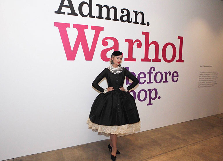 A model dressed in 1050's vintage clothing standing in front of a wall with lettering that reads, Adman. Warhol before pop.