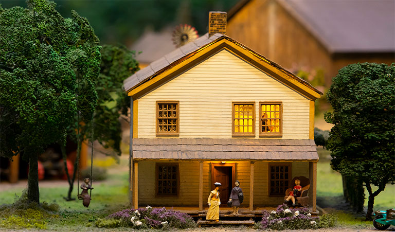 A miniature house with a woman rocking a child on the porch and a woman walking a baby in a upstairs widow of the house.