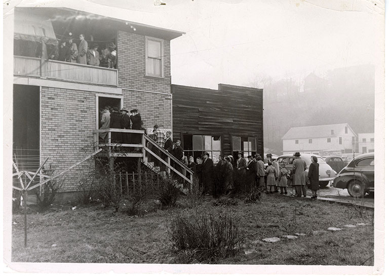 A black and white photo of people lined up wand waiting to get into a house to see a railroad display.
