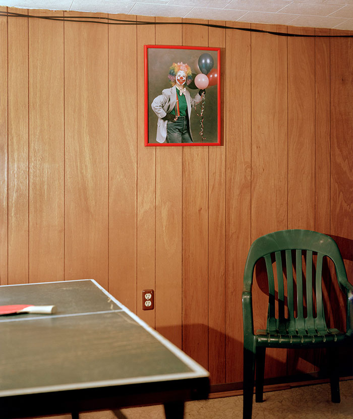 A paneled room with a ping pong table and a painting of a clown hanging on the wall. A green chair sits beneath the painting.