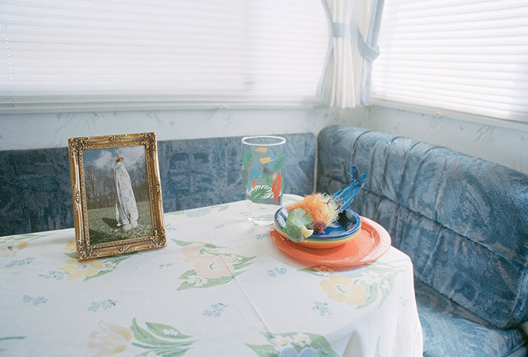 A table setting of a glass, a framed painting of a woman, and a stuffed parrot laying on a stack of colorful dishes.
