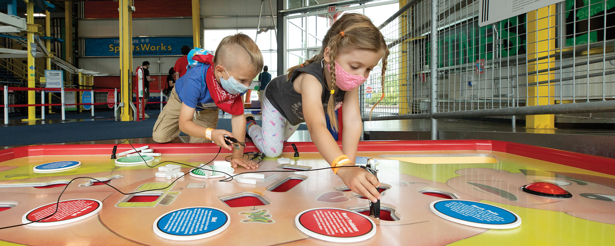 Two children playing on a giant game of Operation. Each child is wearing a mask.