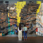 Two people standing in front of a large tapestry made of bottle caps and metal scraps.