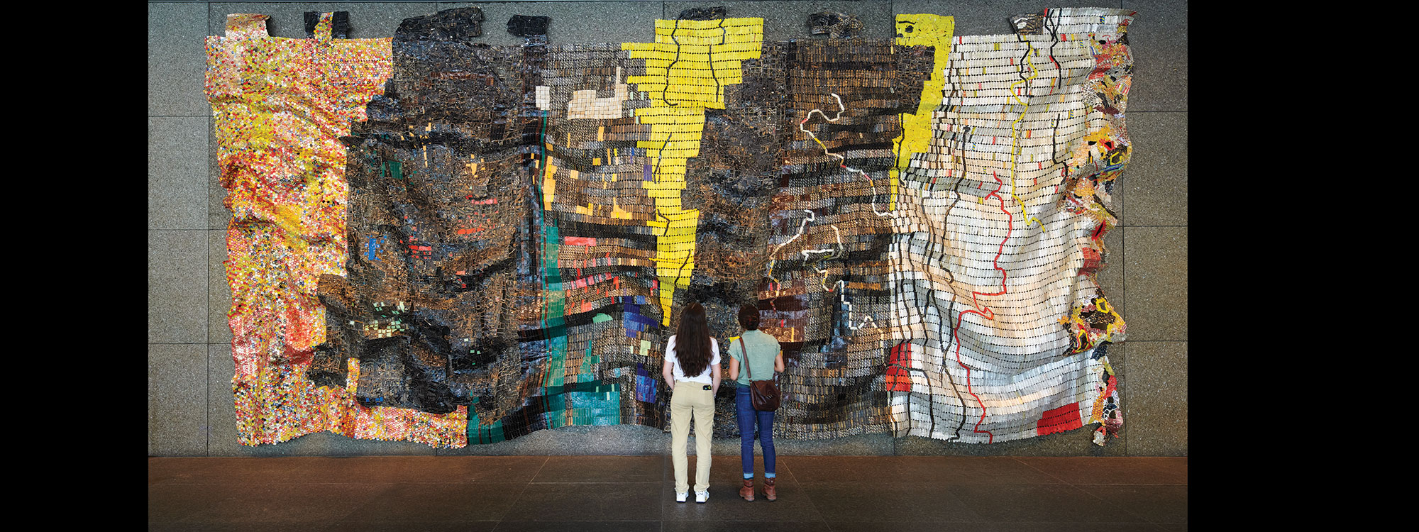 Two people standing in front of a large tapestry made of bottle caps and metal scraps.