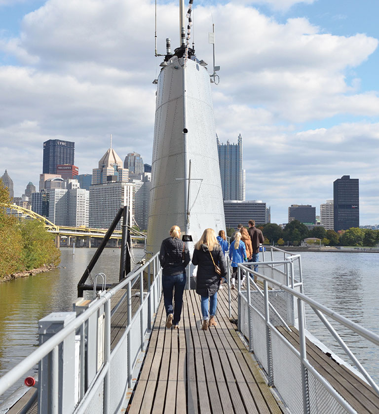 People on the deck of a submarine with the city of Pittsburgh in the background.