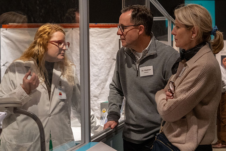 A conservator, in a white lab coat, speaking to museum guests through a glass window.