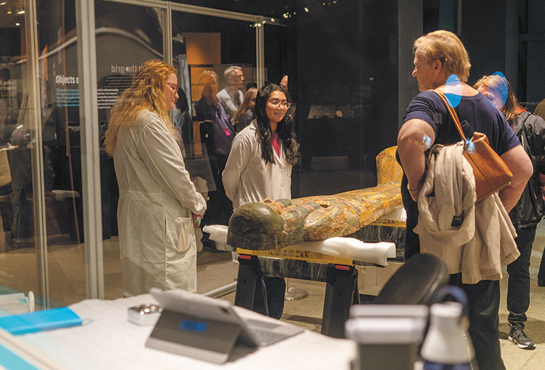 Two museum conservators talking to museum guests standing around a colorful sarcophagus.