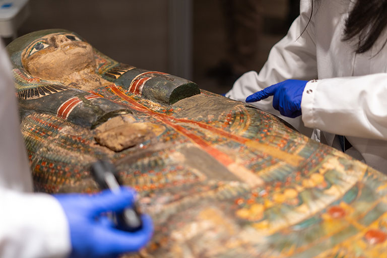 gloved hands of two conservators, working on a colorful ancient egyptian sarcophagus.