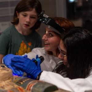 Two museum conservators working on a colorful egyptian sarcophagus, with a young girl standing near them watching.