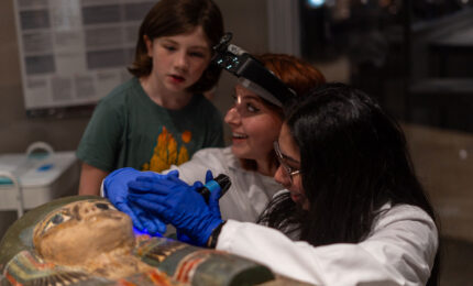 Two museum conservators working on a colorful egyptian sarcophagus, with a young girl standing near them watching.