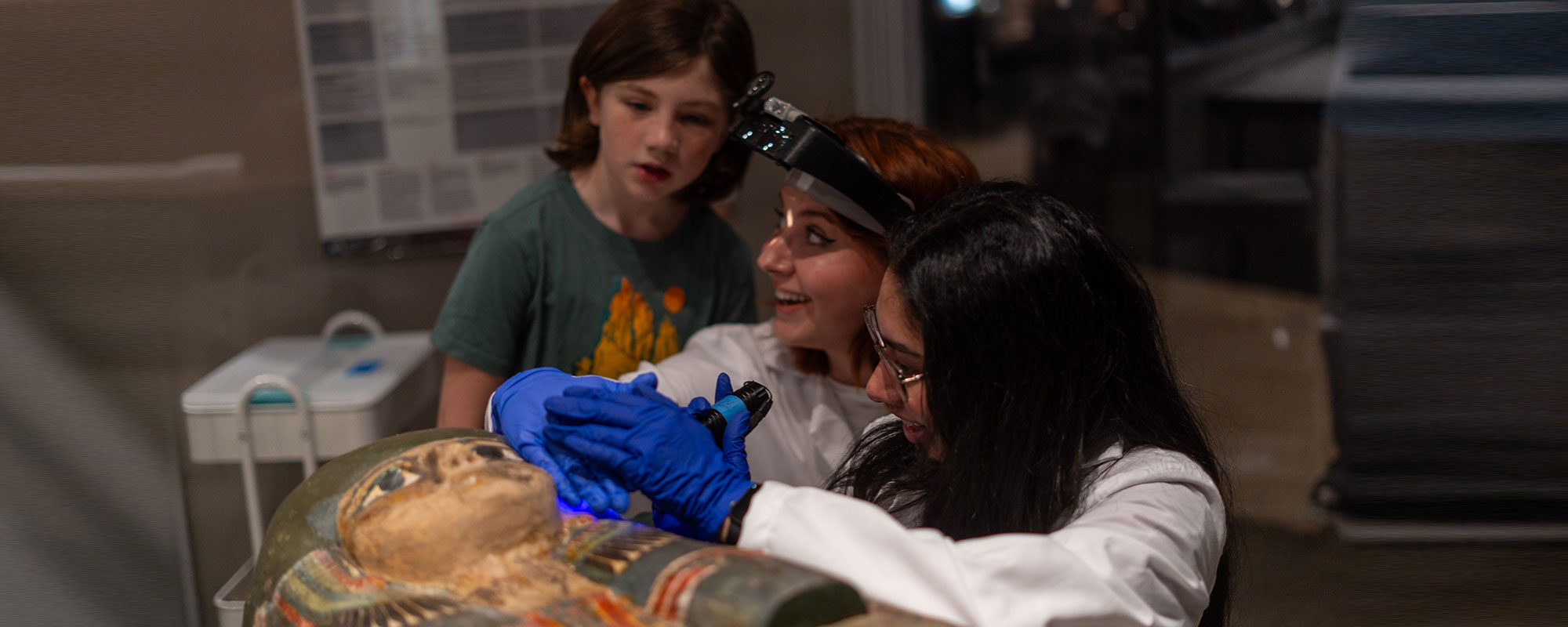 Two museum conservators working on a colorful egyptian sarcophagus, with a young girl standing near them watching.