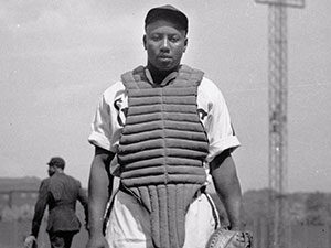 A black and white photo of an Black baseball player wearing a catchers vest.