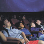 A group of children seated in a planetarium, staring at a star filled screen.
