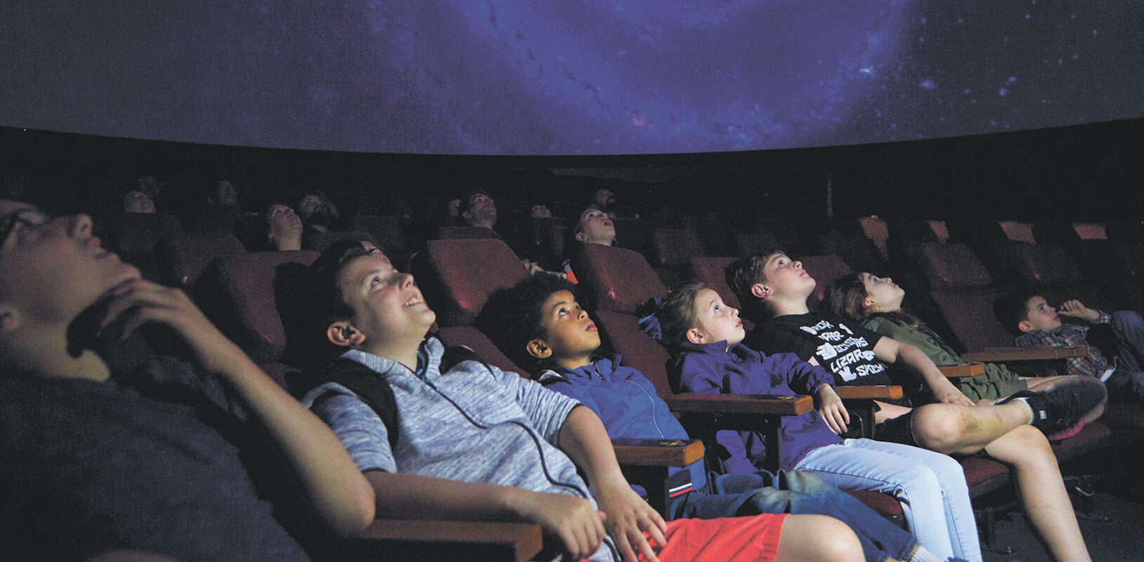 A group of children seated in a planetarium, staring at a star filled screen.