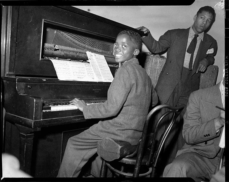 A black and white photo of a young boy playing the piana, and smiling at the camera as two adult men nearby.