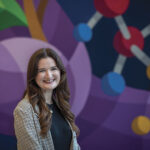 A woman in a plaid blazer smiles in front of a colorful mural depicting molecular structures. The background is vibrant and abstract, creating a cheerful atmosphere.