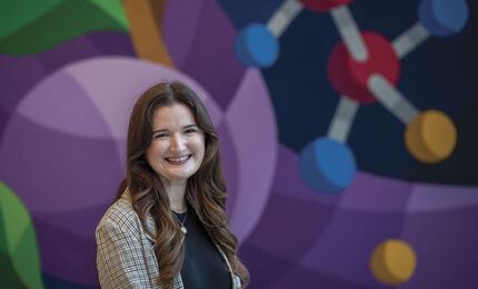 A woman in a plaid blazer smiles in front of a colorful mural depicting molecular structures. The background is vibrant and abstract, creating a cheerful atmosphere.