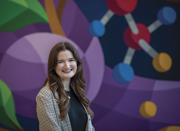 A woman in a plaid blazer smiles in front of a colorful mural depicting molecular structures. The background is vibrant and abstract, creating a cheerful atmosphere.