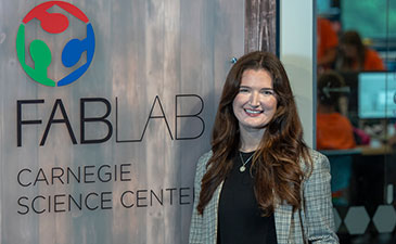 A woman with long brown hair smiles in front of the Fab Lab sign at Carnegie Science Center. The background shows a tech workshop with people working.