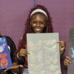 Three smiling individuals hold colorful art pieces against a purple backdrop. The art includes vibrant flowers, butterflies, and pigeons. Energetic and creative atmosphere.