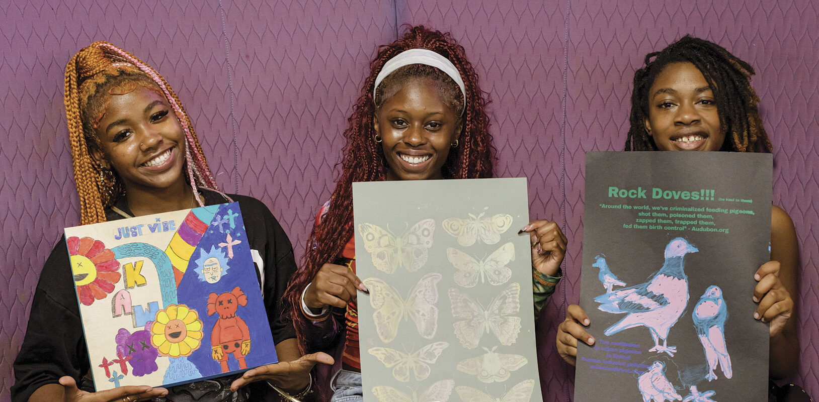 Three smiling individuals hold colorful art pieces against a purple backdrop. The art includes vibrant flowers, butterflies, and pigeons. Energetic and creative atmosphere.