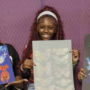 Three smiling individuals hold colorful art pieces against a purple backdrop. The art includes vibrant flowers, butterflies, and pigeons. Energetic and creative atmosphere.