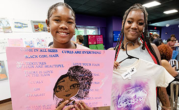 Two smiling girls hold up a pink poster celebrating black hair and a T-shirt. The room has colorful artwork in the background, creating a joyful atmosphere.
