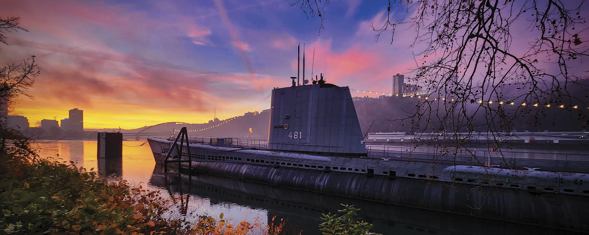 A submarine docked by a riverbank under a vibrant purple and orange sunset. Silhouetted city buildings and a bridge are visible in the background.