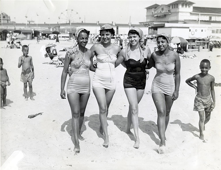 Four women in vintage swimsuits walk arm-in-arm on a lively beach. They are smiling, conveying a joyful and relaxed atmosphere. Ferris wheel and people in background.