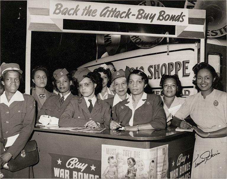 Group of women in military and civilian attire pose at a "Buy War Bonds" booth with signs urging support. The tone is serious and determined.