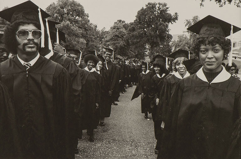 Graduates in caps and gowns stand in two lines on a tree-lined street, smiling and celebrating graduation, conveying joy and achievement.